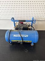 Blue Mastercraft air compressor tank viewed from an angle showing pressure gauges, black hose, and handle.