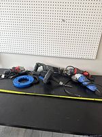 Power tools including a cordless reciprocating saw, drain snake drum, and power tool attachment on a black table under pegboard