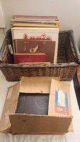 Wicker basket holding multiple vinyl records including an original motion picture soundtrack titled 'Amos'. Below it is a worn cardboard box containing more records.