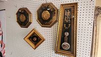 Set of four Roman-themed wall hangings displayed on a pegboard wall, chains and hooks visible for hanging. One tall rectangular piece and three smaller decorative framed pieces, all gold and black with relief and painted details.