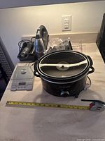 Photo showing black Crock-Pot slow cooker with glass lid, handheld mixer and digital scale on kitchen counter with measuring tape for scale