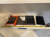 Photo showing the lot with an Amazon Fire tablet box, Acer tablet with case, and Kobo e-reader with case placed in a white shelving unit with a measuring tape.