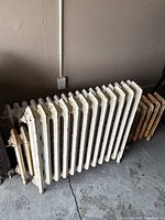 Side angle view showing two white radiators and one rusted radiator all resting on a concrete floor against a wall with an electrical outlet.
