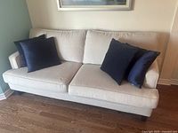 Front view of beige fabric two-seat sofa with two dark blue throw pillows on a brown wooden floor against a white wall.