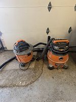 Photo of both Ridgid wet/dry shop vacuums side by side on garage floor with hoses and power cords visible, showing overall condition and size comparison.