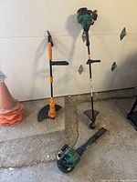 Photo showing two whipper snippers standing upright and one leaf blower laying on the ground in a garage setting.