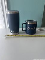Both navy blue Yeti travel mugs on white surface with measuring tape showing sizes, one is taller cylindrical tumbler and one shorter with handle and designs.