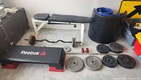 Weight bench, barbell with attached weights, multiple weight plates, red collars, black mats, and Reebok step platform shown from front view