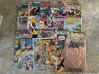 Twenty Avengers comic books spread out in three rows on carpet. Covers visible showing a mix of action scenes and characters from Marvel's Avengers series.