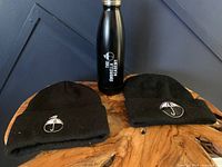 Photo of two black knit toques and one black water bottle with Umbrella Academy logo on round wooden surface against dark background.