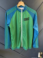 Front view of green and blue zip-up jacket with FUBAR 2 logo on left chest, hanging on hanger.