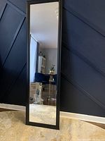 Full-length black framed mirror positioned upright against a navy blue paneled wall, reflecting a part of the room including white curtains, a dark blue chair, and a small side table