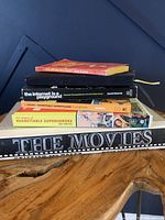 Stack of five books on a wooden table: 'The Movies', two copies of 'The Issues of Regrettable Superheroes', 'The Internet is a Playground', and a small red book on top.