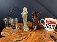 Lot displayed on wooden surface with a blue background showing 4 clear glass Leslieville Beer Company drinking cups, 2 statues (frosted glass and dark brown figurine), and a white ceramic coffee mug with text