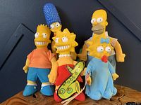 Group photo showing all five Simpsons plush dolls standing together; Homer, Bart, Lisa, Maggie with tags, Marge without tag