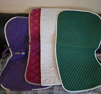 Four assorted quilted equestrian saddle blankets in purple, red, white, and green colors laid out side by side showing pattern and edges.