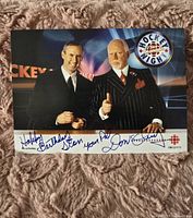 Front view of a signed color photo featuring Don Cherry and another man standing in front of a Hockey Night in Canada backdrop, with inscriptions and signatures below the photo.