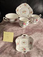 Image of Royal Albert Petit Point china pieces arranged on a cloth showing two-handled sugar bowl, two teacups, and two square plates with floral patterns.