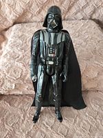 Black 15-inch Darth Vader action figure from Star Wars with a fabric cape, standing upright on a textured pink surface.