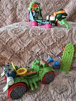Two TMNT toy vehicles displayed together. The green Toilet Taxi vehicle with toilet seat in front, and the Skateboardin' Mike vehicle behind with attached figure and pink wheels.