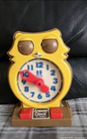 Front view of the yellow owl-shaped plastic clock toy showing the white dial with blue numbers and red hands pointing at 10:25. Toy has molded owl features and red buttons at the base.