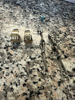 Photo shows two pairs of earrings and one ring arranged on granite surface. The silver braided ring stud earrings with chains and beads, a pair of gold-tone floral etched earrings, and a silver ring with turquoise stone are clearly visible.