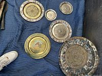 Top-down photo of 6 decorative metal circular pieces featuring Aztec/Mayan calendar designs in various sizes. Some are brass, others silver-tone with engraved details and one has colorful Aztec artwork.