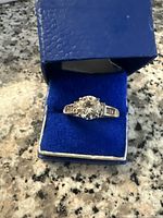 Close-up image of the silver ring with central round gemstone displayed in the blue velvet ring box on a granite surface.