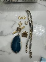 Jewelry lot on marble surface including two pairs of gold-toned textured earrings, a two-tone metal chain necklace, ornate gold-tone cross pendant, blue stone pendant, and small textured charms.