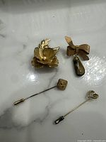 Photo showing gold-plated flower brooch and three stick pins with pearl and decorative designs on a white marble-like surface.