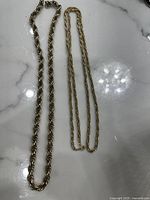 Two gold rope chain necklaces laid flat on a white surface, showing the difference in thickness and design of the chains.