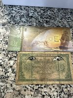 Gold bar in original packaging alongside a 10 Belgian Francs banknote on a granite surface.