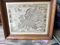 Framed artwork showing an antique-style map of Europe with detailed border illustrations and wooden frame.