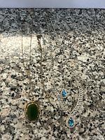 Three vintage style jewelry necklaces placed on granite surface: gold-tone with large green stone, silver-tone with blue and clear stones, silver-tone chain with small crucifix pendant