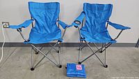 Two blue folding camping chairs side by side with a folded chair and carrying bag at their base on a concrete floor.