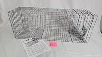 Full view of metal wire humane animal trap with single door, laid on white background with instruction sheet nearby.