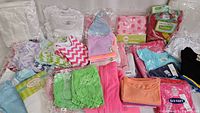 Wide view of assorted new baby and toddler clothing including bibs, shorts, onesies, gowns, and blankets with various colors and patterns
