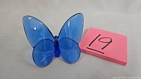 Front view of blue crystal butterfly figurine next to pink paper with number 19, showing overall shape and color.