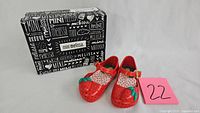 Pair of red Mini Melissa children's shoes with strawberry embellishments next to original branded box
