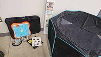 Photo showing folded black pet playpen with turquoise trim, portable carrying bag, interactive puzzle toy, and instruction manual on floor.