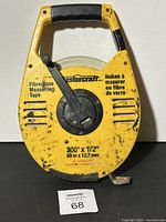 Yellow MasterCraft fiberglass measuring tape with black crank handle and rubber grip, showing front view of case with printed measurement details and minor wear marks.