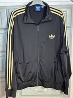 Front view of black Adidas jacket with gold stripes running along the sleeves and Adidas trefoil logo on the chest, displayed on a hanger.