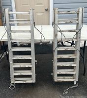 Front view of the two aluminum loading ramps standing upright showing the rungs, securing cables, and safety chains.