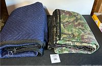 Two folded moving blankets on display, one blue quilted and one camouflage.