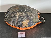 Top view of the round outdoor cushion showing camouflage pattern with orange trim and carabiner attached to black straps