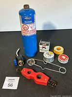 Overview of the entire lot on black surface against white background showing propane tank, Teflon tape rolls, solder wire spools, pipe wrench, and other plumbing tools.