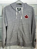 Front view of the grey Roots zip-up hoodie with hood up and visible red maple leaf patch on chest and front pockets