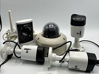 Five security cameras grouped together: Three white Lorex LBV1521-C bullet cameras, one beige Digital Watchdog V4363D dome camera, and one white D-Link DCS-2330L indoor wireless camera with antenna.