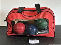Red carrying bag with black handles containing visible blue and red bocce balls, with Sportcraft logo patch.