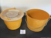 Two vintage yellow minnow buckets side by side on a black surface, one bucket with lid and handle, the other open with metal handle.